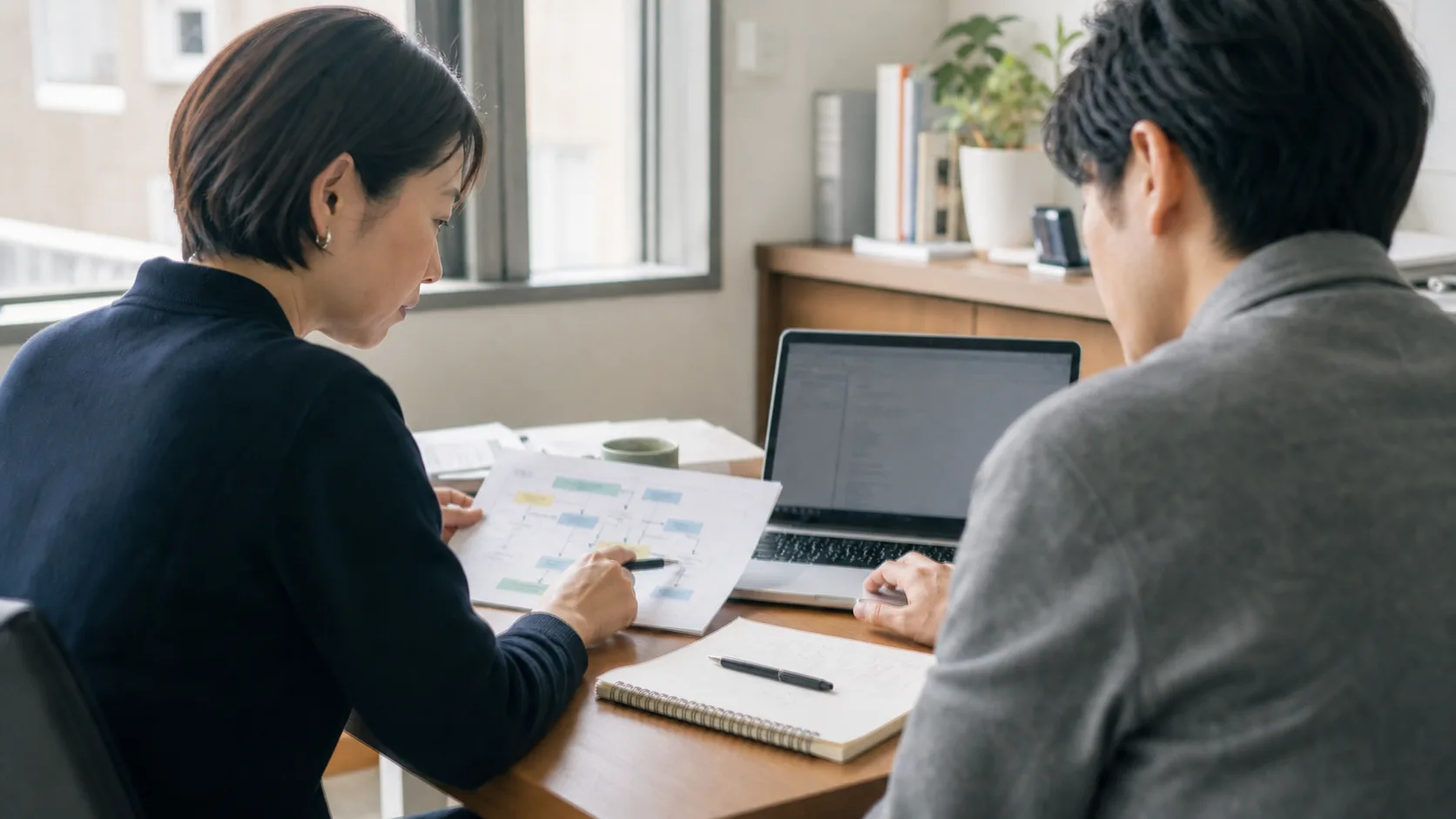 2人が業務の流れを示す資料とノートパソコンを見比べ、人とAIの役割を検討している様子