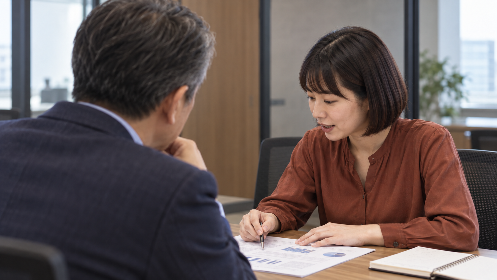 管理職の肩越しに、資料を見ながら話す女性を捉えた対話の場面。